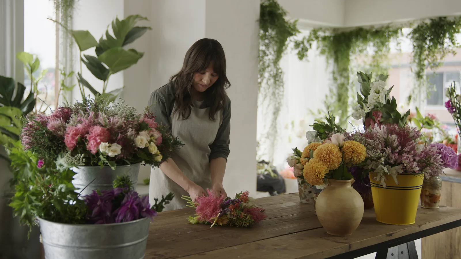 Šviesus gėlių salonas, kuriame floristė prie medinio darbastalio riša puokštę, aplink vazose šviežios gėlės