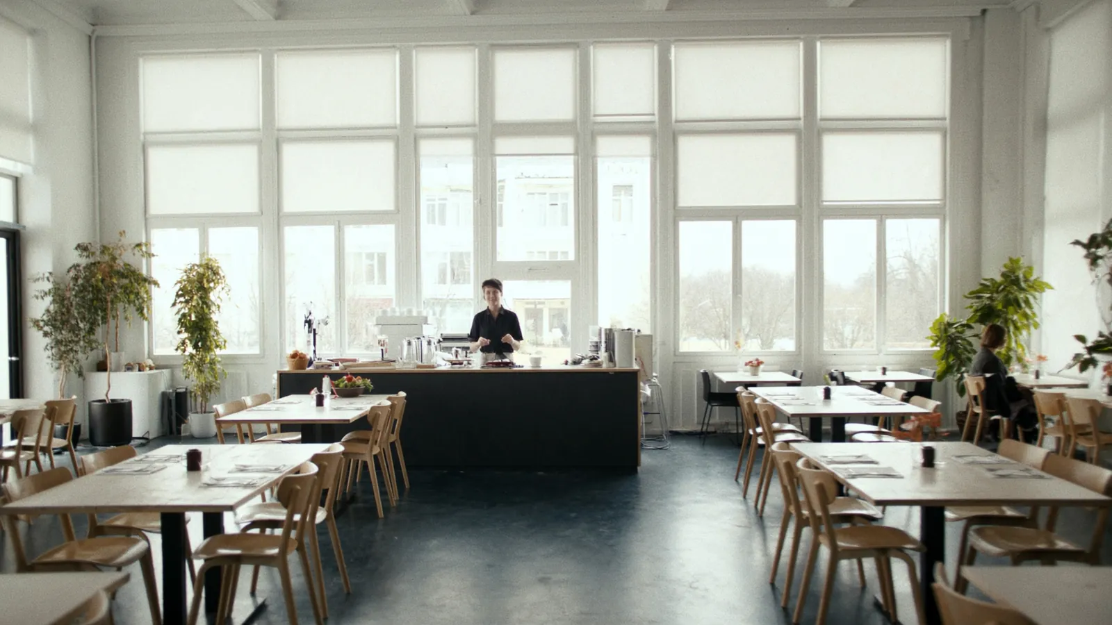 Jauki modernaus restorano ir kavinės vidaus erdvė su mediniais staliukais, šilta apšvietimu ir baristu prie baro